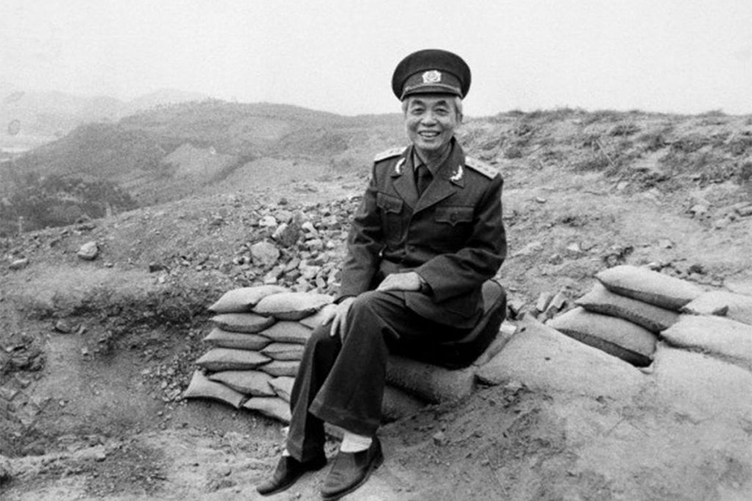 An older man in a military uniform sits on sandbags, smiling, with rocky hills and a barren landscape in the background.