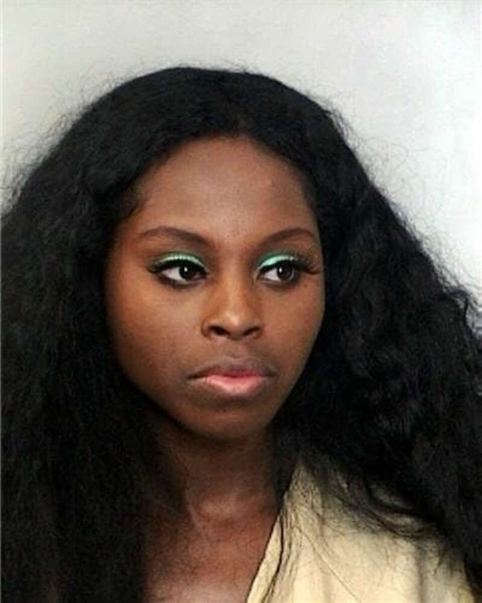 A woman with long, wavy black hair and bright green eyeshadow looks slightly to the side with a neutral expression against a plain, light-colored background.