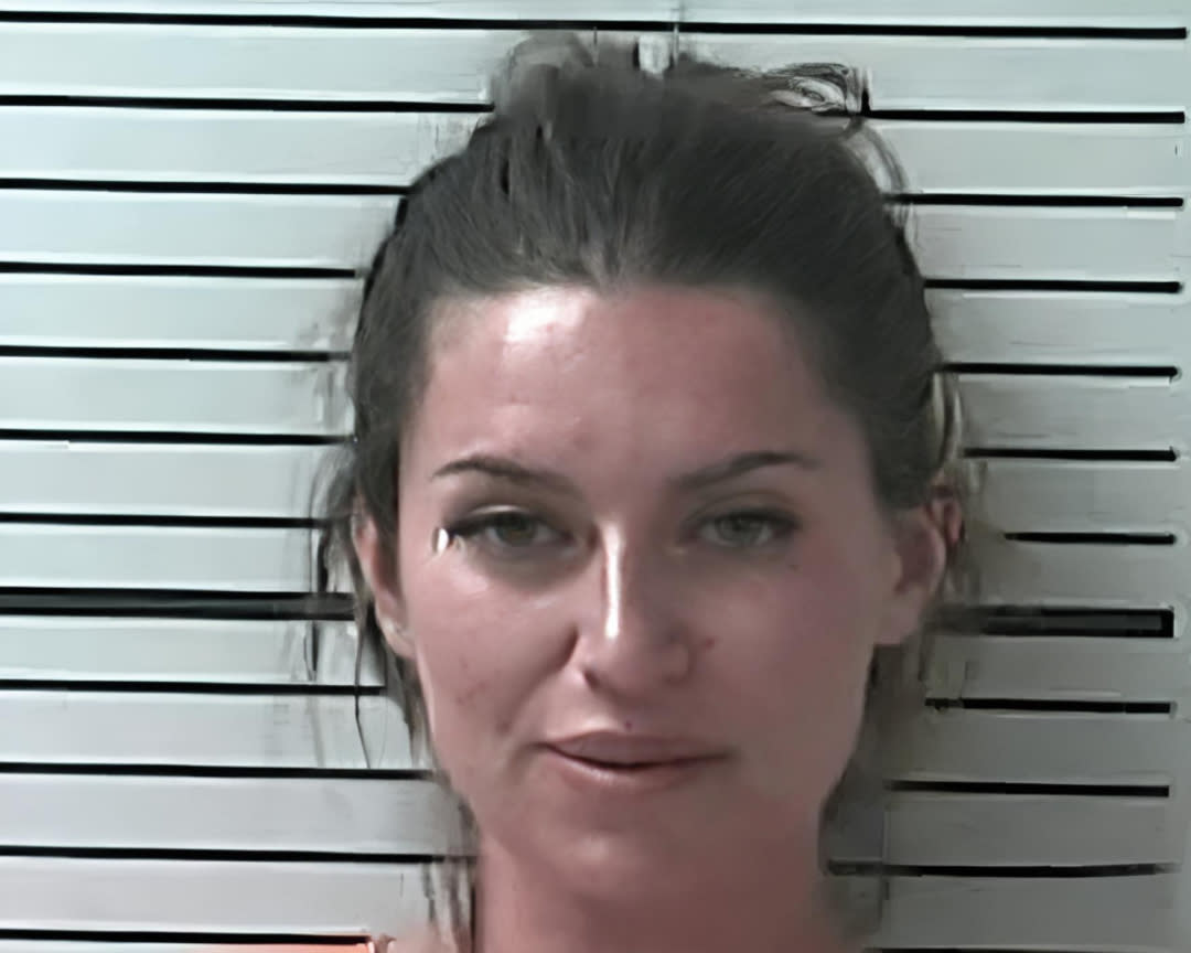 A woman with dark hair pulled back is posing against a height chart with horizontal black lines on a white background, as in a mugshot photo. She is wearing an orange top and looking straight at the camera.