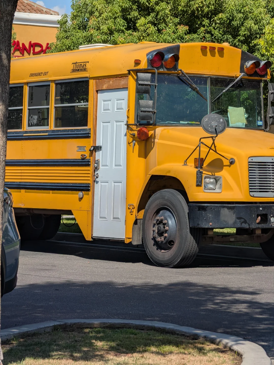 A yellow school bus with a white residential door instead of a standard bus door is parked on a street near some trees and a building.