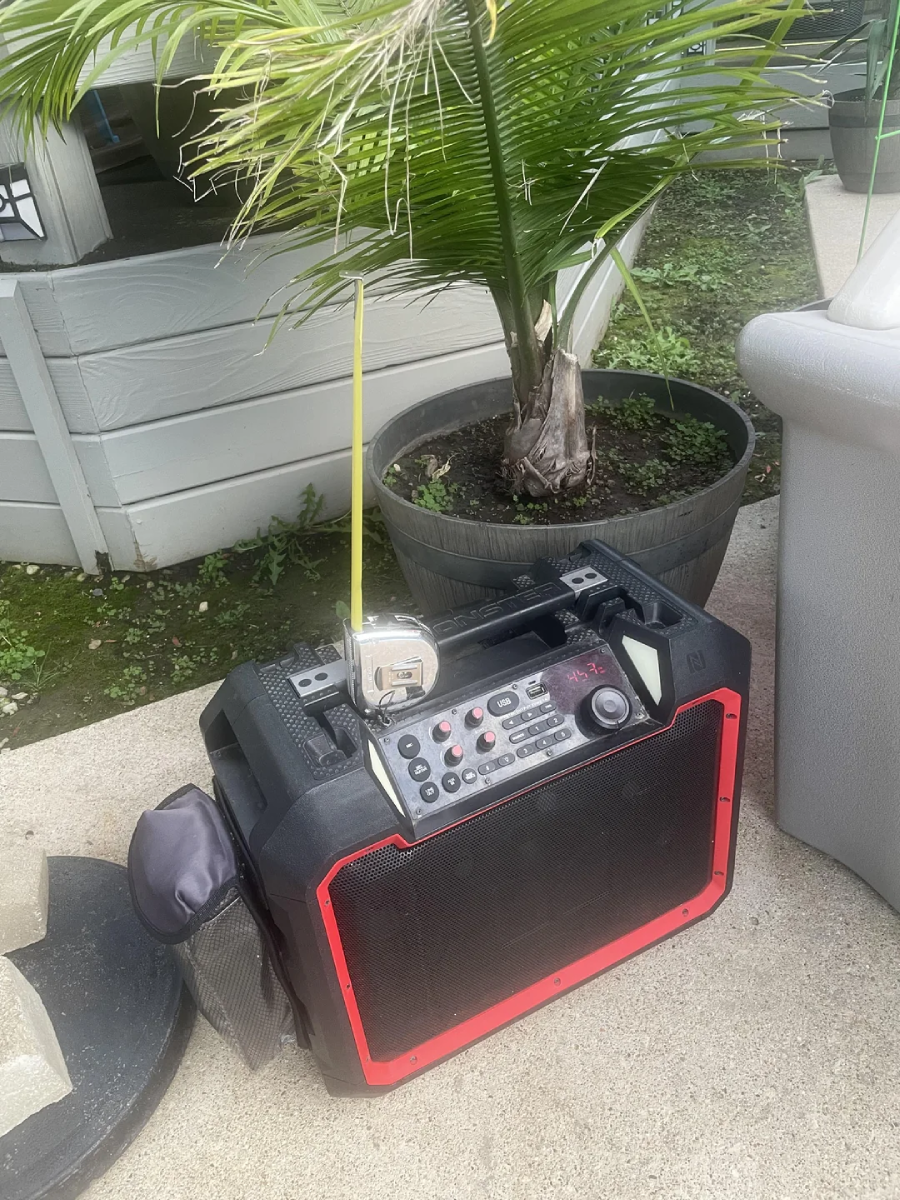 A portable black and red speaker with control buttons sits on a concrete surface next to a potted palm plant and a gray outdoor structure. There is a small electronic device attached to the top of the speaker.
