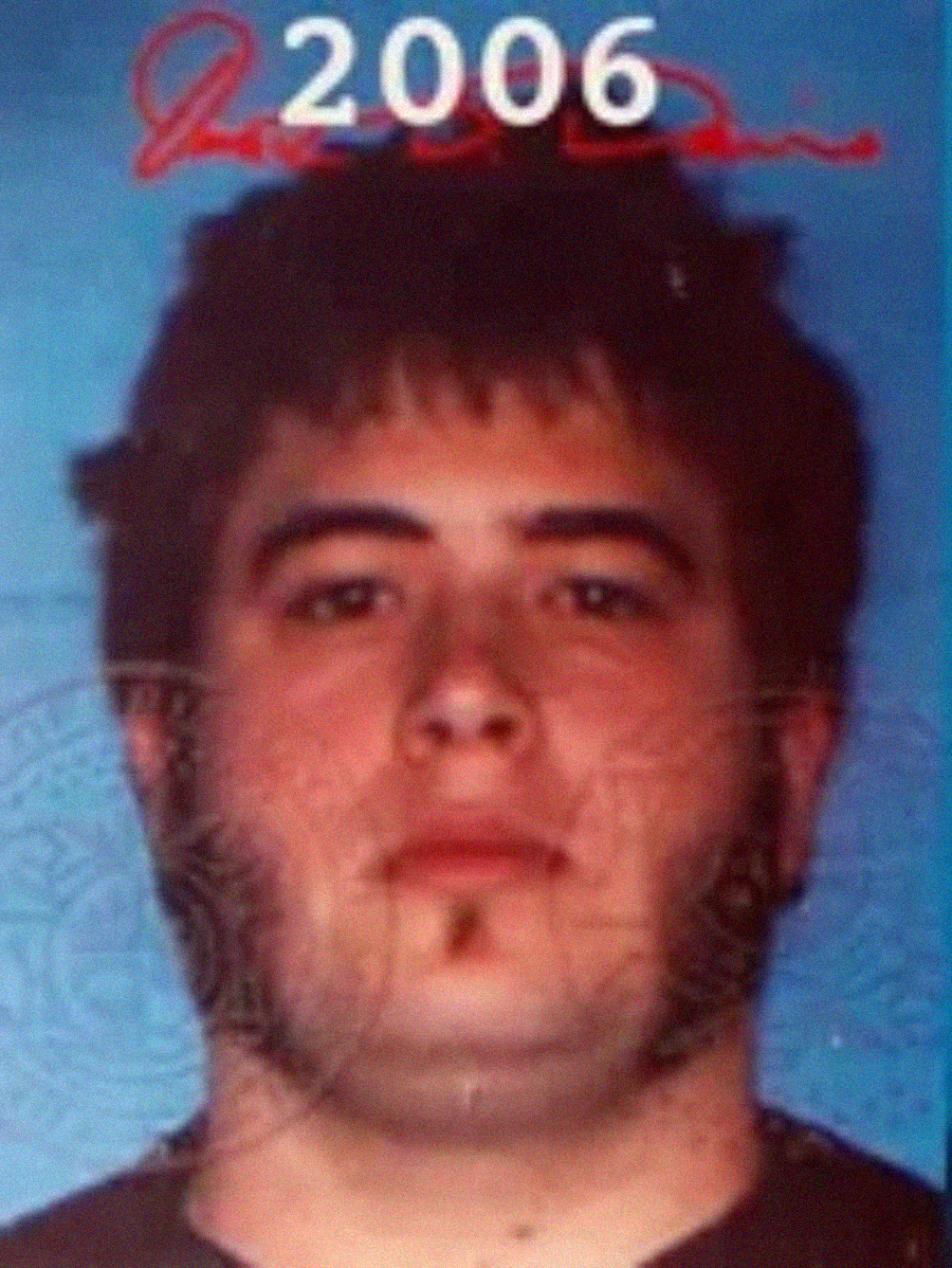 A young man with dark hair, sideburns, and a small patch of facial hair on his chin poses for a photo ID. The year "2006" is visible at the top of the image. The background is blue.
