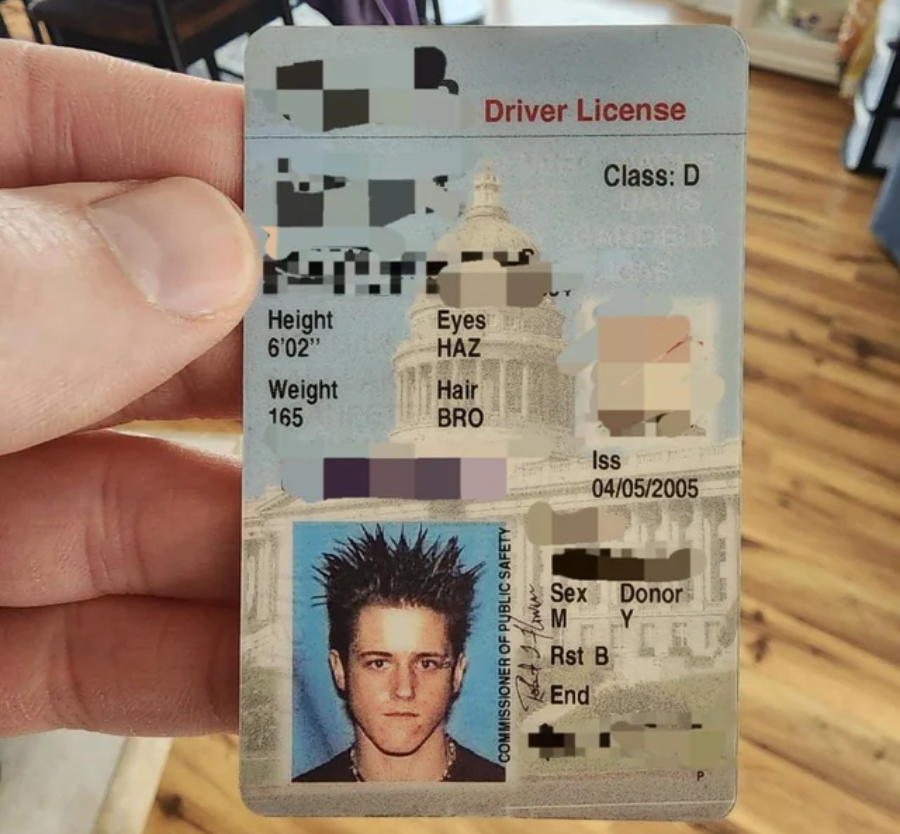 A hand holds up a driver license displaying a person with spiked dark hair in the photo. Parts of the personal information are obscured. The background includes a building with columns and wooden flooring.