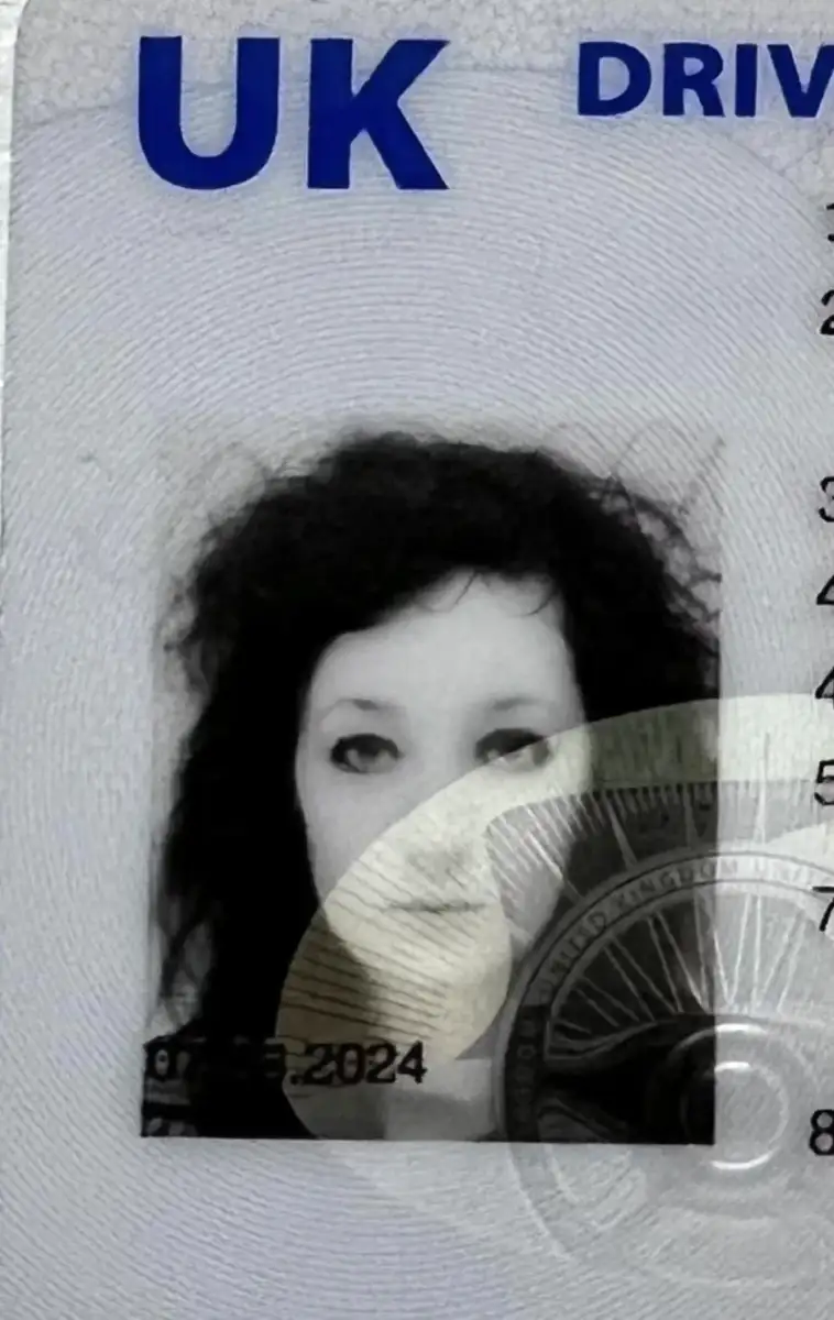 A close-up of a UK driving licence showing a black-and-white ID photo of a person with curly dark hair and light skin. The top of the card displays bold blue "UK" text. A security hologram partially covers the photo.