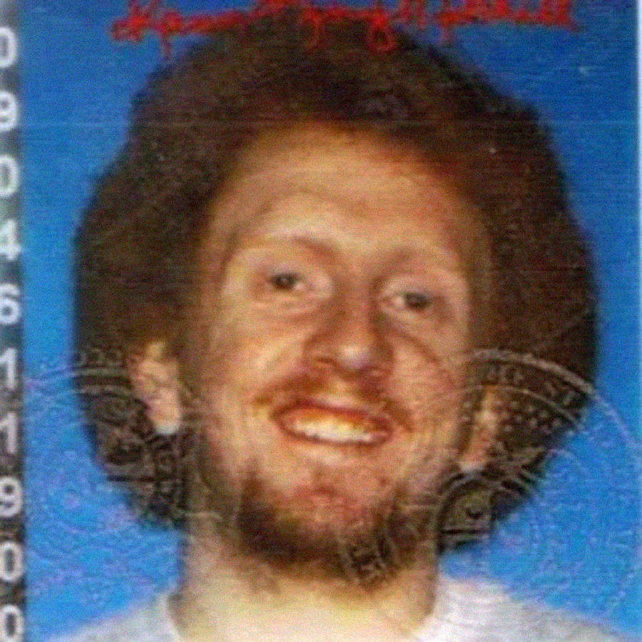 A man with curly brown hair and a beard smiles at the camera against a blue background, as seen in a photo ID with numbers visible along the left edge and a faint security seal across the image.