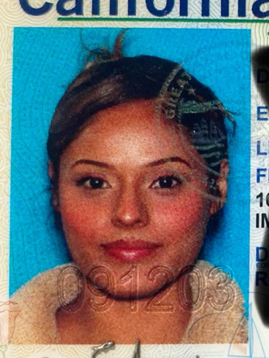A woman with light brown skin and brown hair pulled back is shown in a close-up ID photo with a blue background and visible official markings.