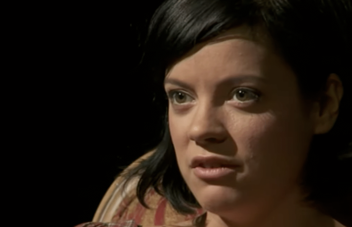 A woman with short dark hair and light eyes sits in a chair, looking slightly to the side with a serious expression. The background is dark, focusing attention on her face.