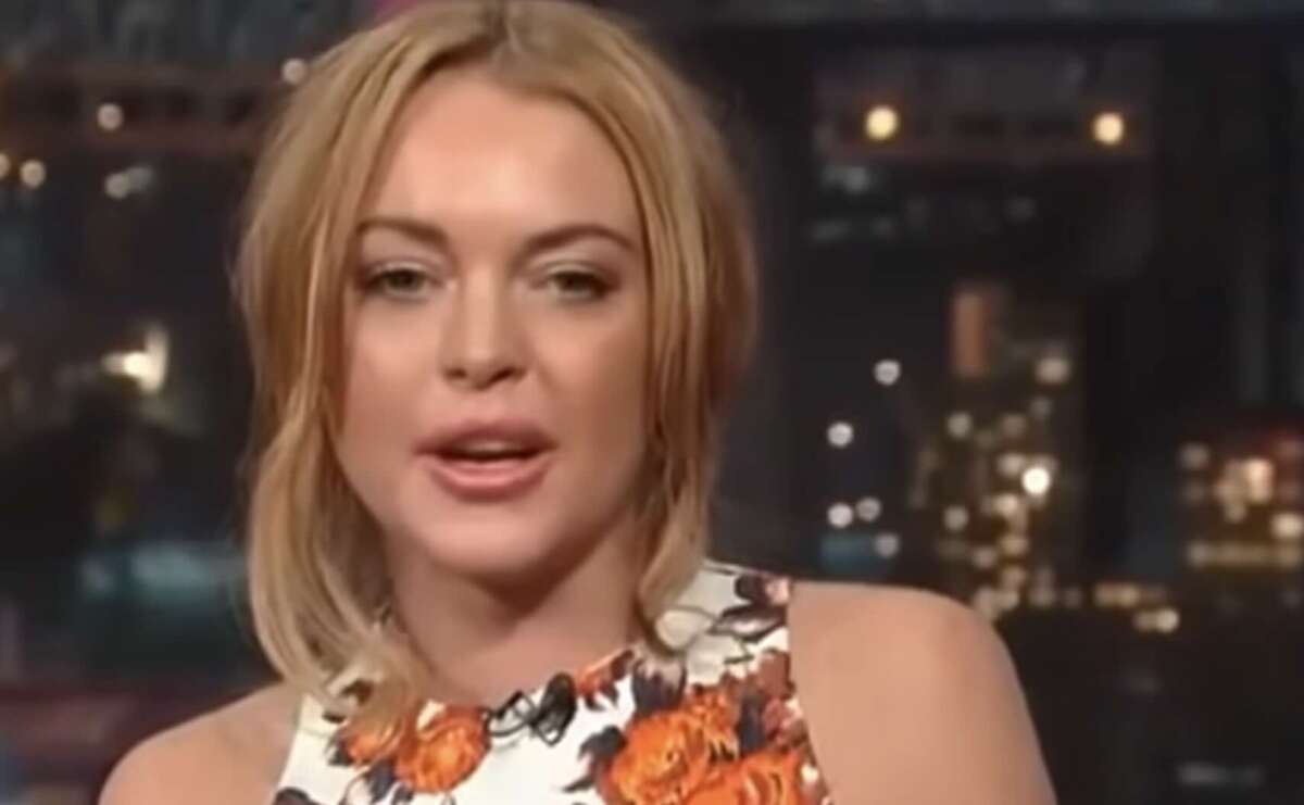 A woman with blonde hair and a floral top is speaking on a talk show set, with blurred city lights in the background.