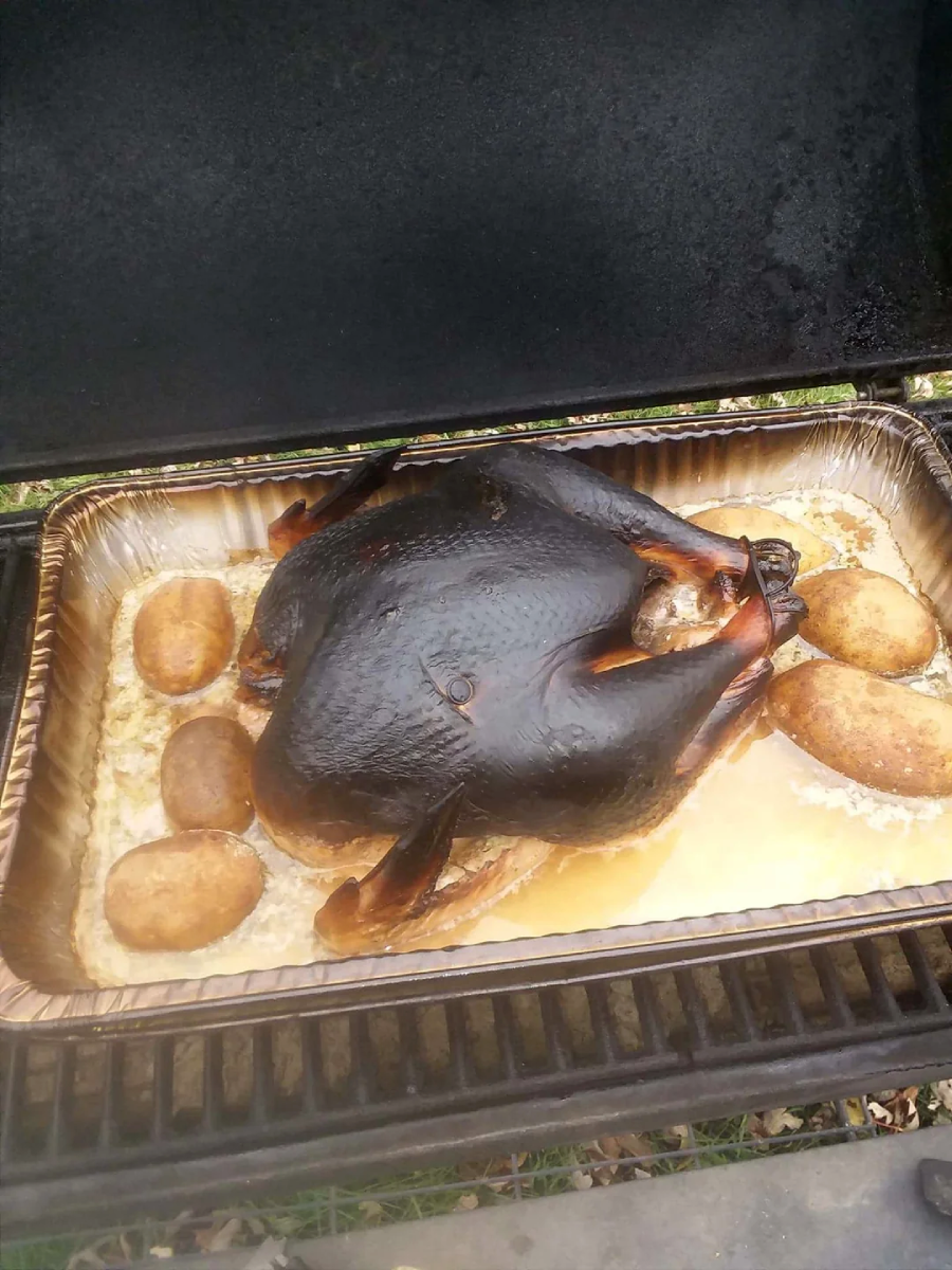 A whole turkey with severely burnt, blackened skin sits in a roasting pan surrounded by several baked potatoes, all inside a grill or smoker.
