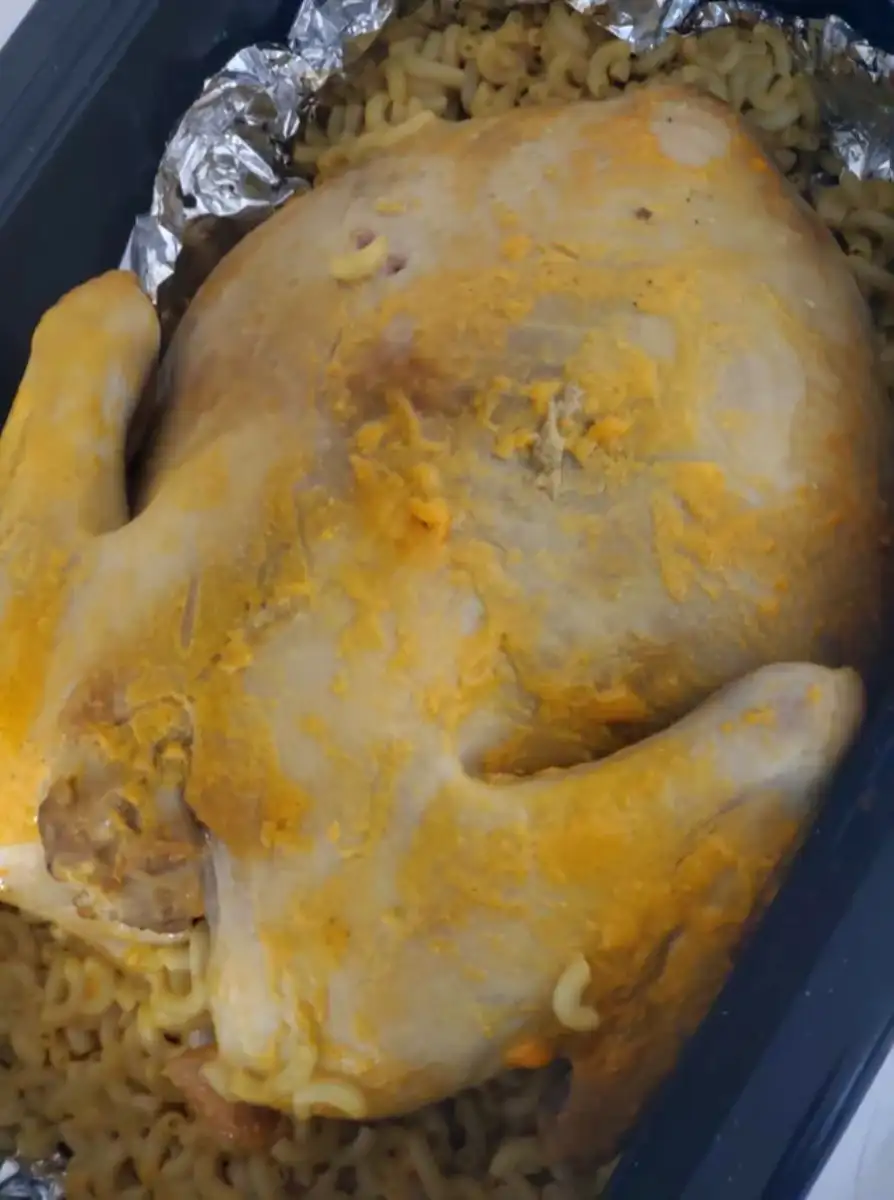A whole cooked chicken covered in yellow seasoning rests on a bed of curly pasta noodles, all placed on aluminum foil in a black rectangular baking tray.