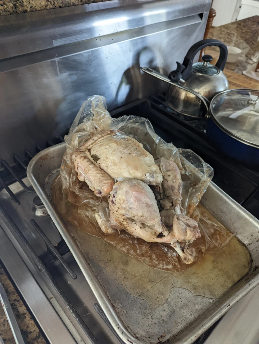 A cooked turkey sits in a roasting pan inside a clear oven bag on a stovetop, with a kettle and a covered pot in the background.