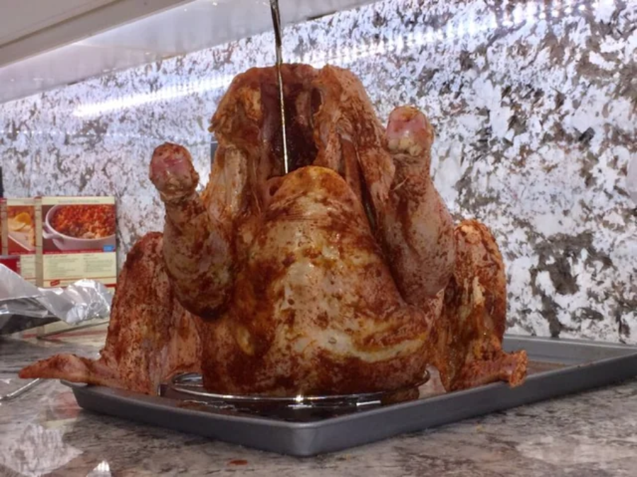 A raw, seasoned whole turkey sits upright on a baking tray in a kitchen, with a dark liquid being poured inside it. Recipe boxes and a marble-patterned backsplash are visible in the background.