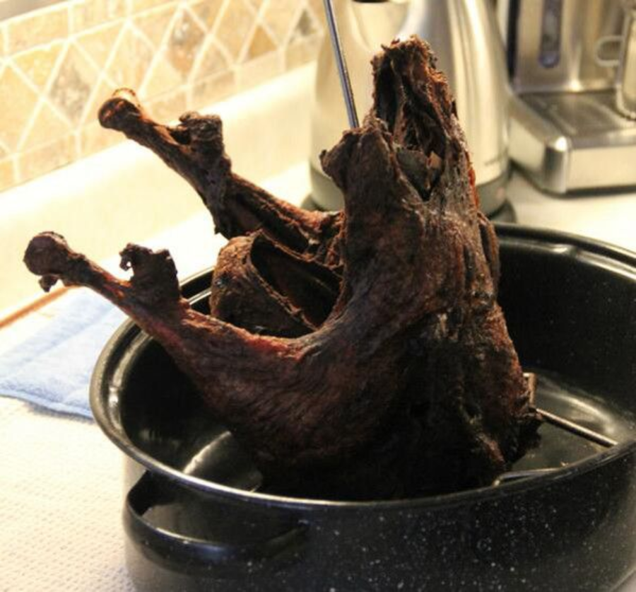 A cooked, darkly roasted turkey is propped upright on a metal spit in a black roasting pan on a kitchen counter, with kitchen appliances and a tiled backsplash in the background.