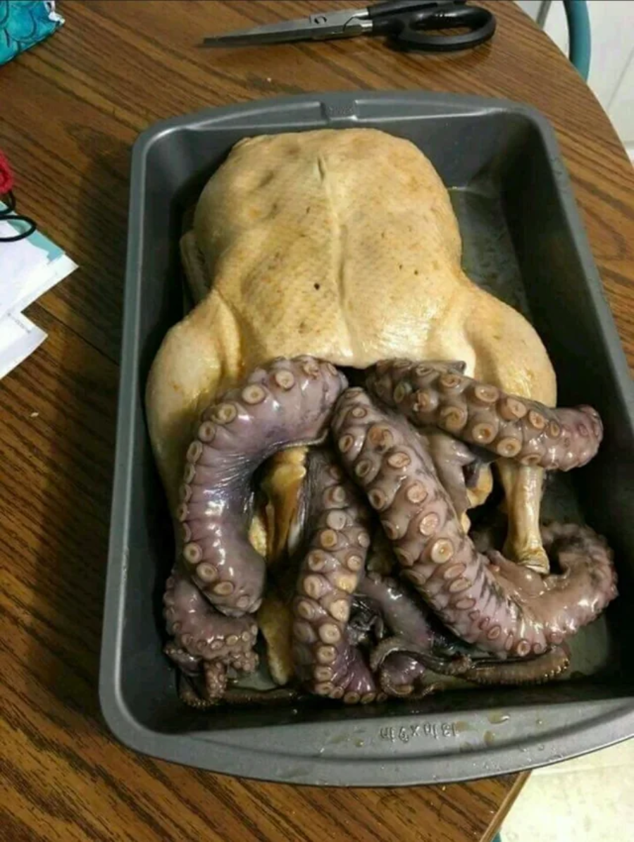 A raw whole chicken in a baking pan with large octopus tentacles spilling out from its cavity, on a wooden table with scissors and papers nearby.