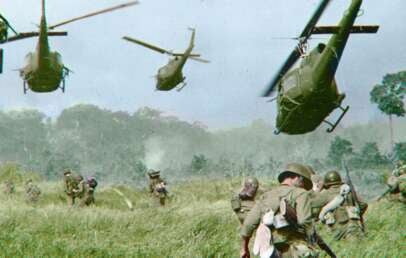 Soldiers move through tall grass as several military helicopters hover above, preparing to land in a jungle clearing. The scene suggests a tense, active combat or military operation.