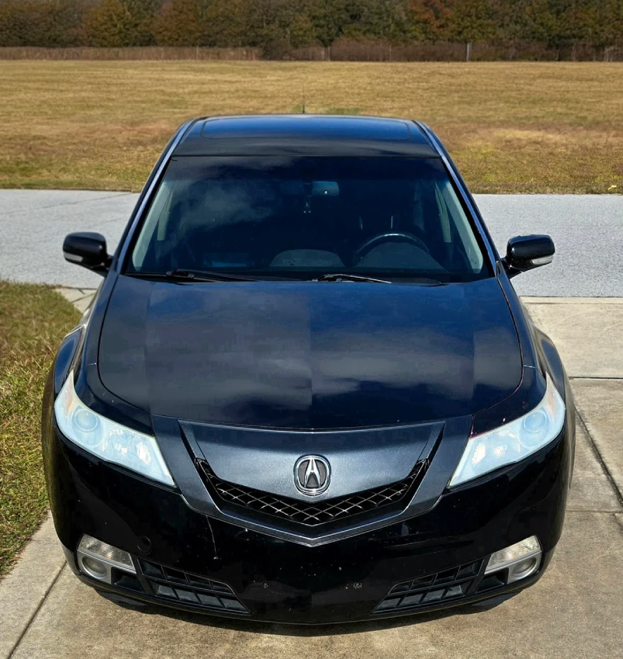 A black Acura sedan is parked on a concrete driveway with grass and an open field in the background, viewed from the front.