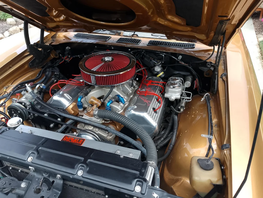 A close-up view of a car engine bay with the hood open, featuring a large V8 engine with a red air filter and red ignition wires inside a gold-painted classic car.