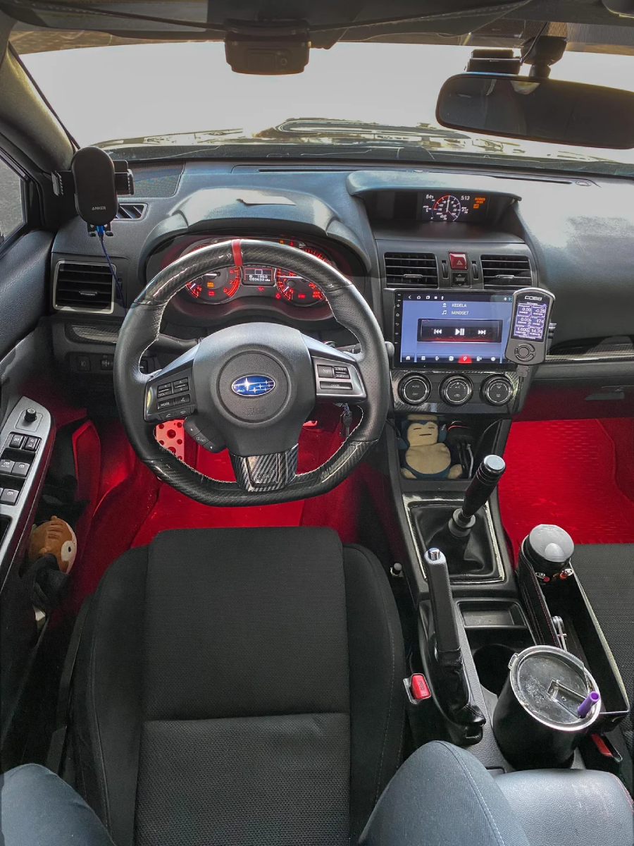 Interior view of a Subaru car showing the steering wheel with the Subaru logo, dashboard with red accents, touch screen display, manual gear shift, cup holder with a drink, and various controls.