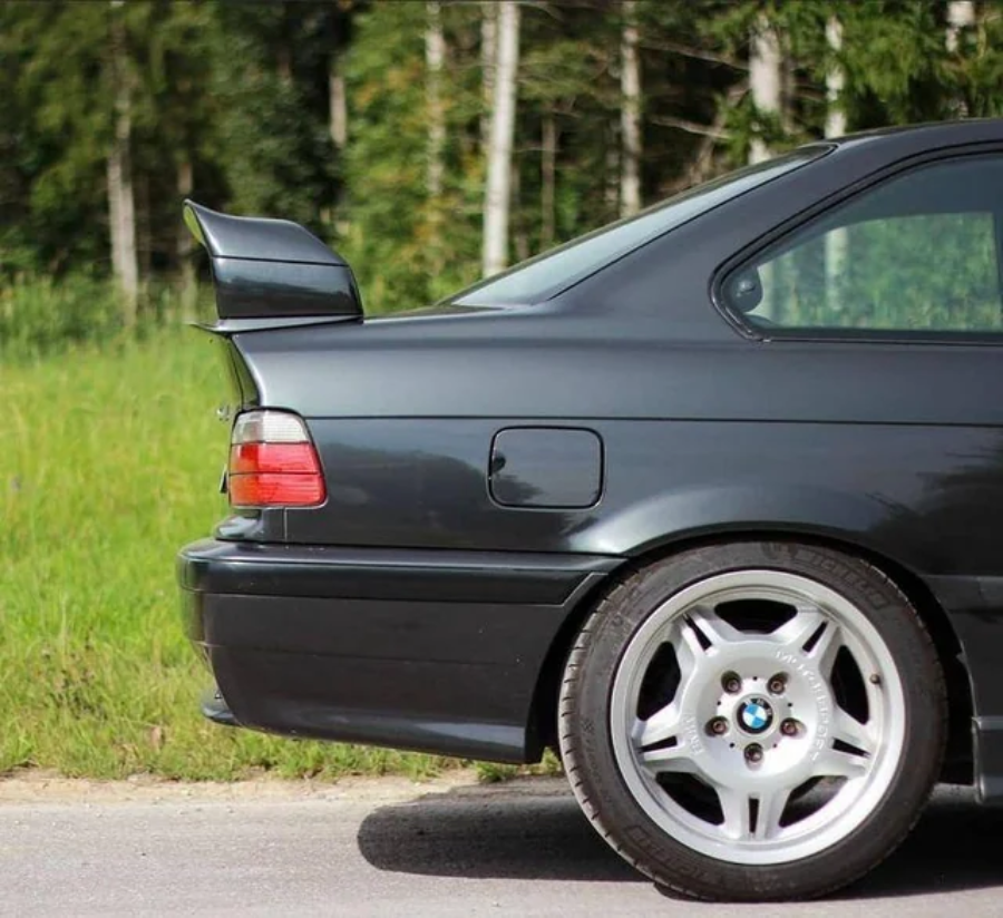 The rear side of a dark-colored BMW car with a rear spoiler is shown parked on a road, with green grass and trees in the background.