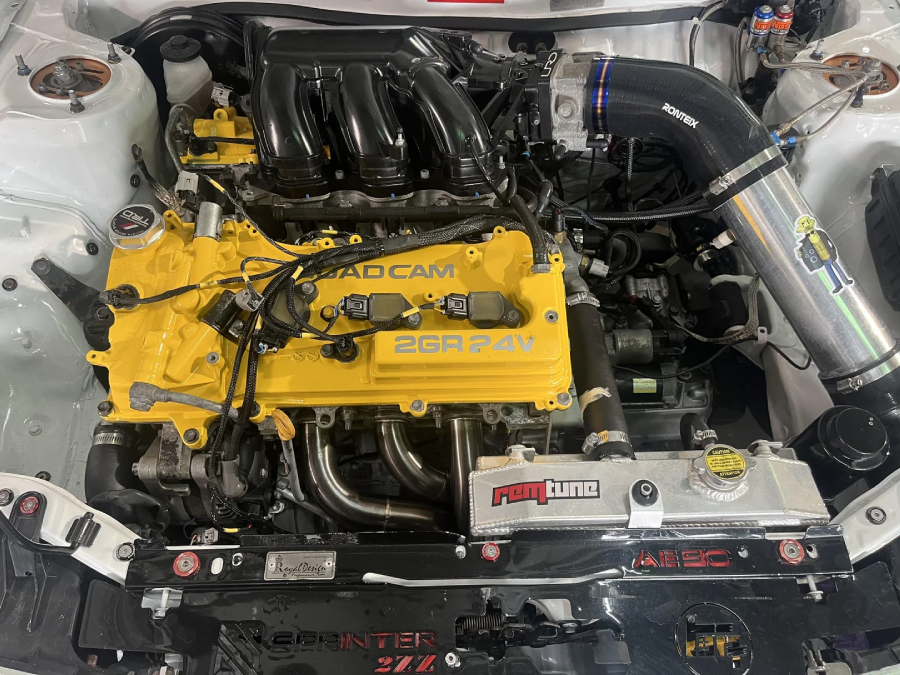 Close-up of a car engine bay with a yellow valve cover labeled "BAD CAM 2GR 24V," visible intake manifold, custom air intake, Ron Tune radiator, and various performance car parts installed.