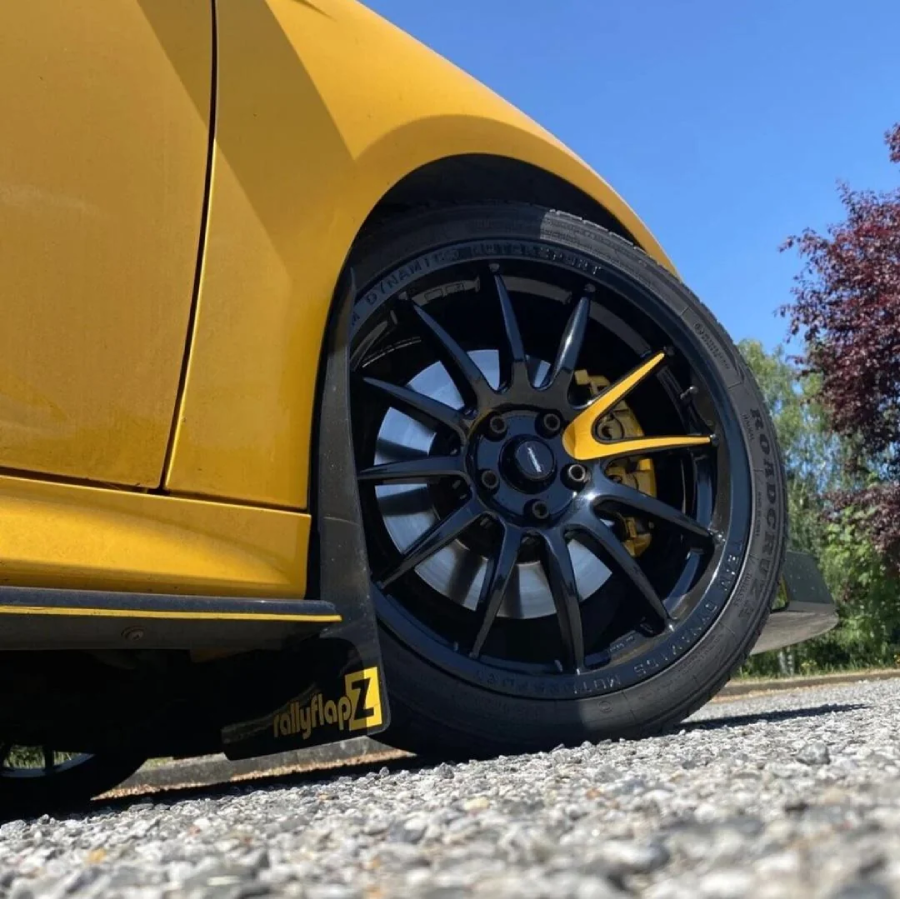 Close-up of a black alloy wheel and tire on a yellow car, parked on a gravel surface. The car has a "rallyflapZ" mud flap, and trees are visible in the background under a clear blue sky.