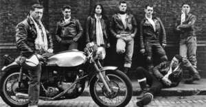 A group of six men and one woman in leather jackets pose against a brick wall; one man sits on a classic motorcycle while another sits on the curb. The overall scene has a vintage, rebellious style.