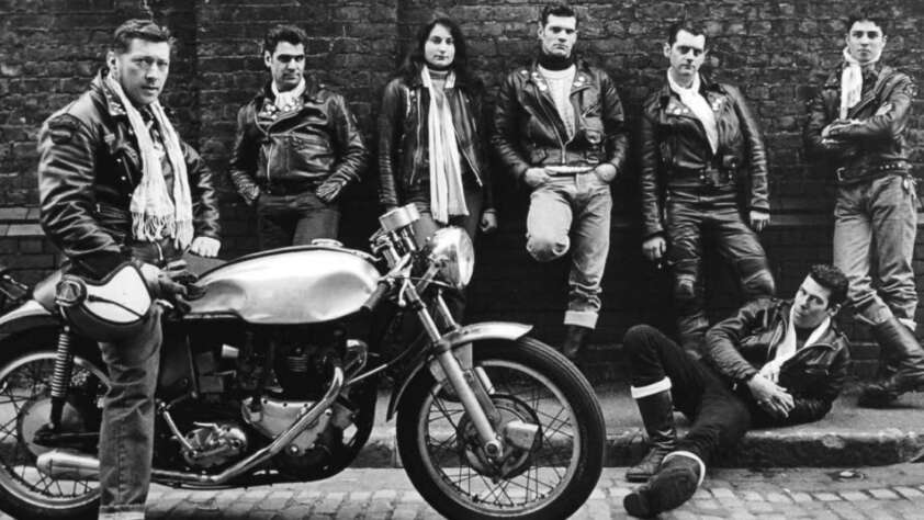A group of six men and one woman in leather jackets pose against a brick wall; one man sits on a classic motorcycle while another sits on the curb. The overall scene has a vintage, rebellious style.