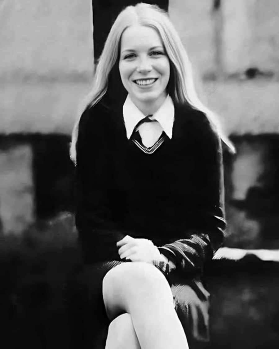 A young woman with long, straight hair sits outdoors, smiling at the camera. She is wearing a dark sweater, white collared shirt, and skirt, with her legs crossed. The photo is black and white.