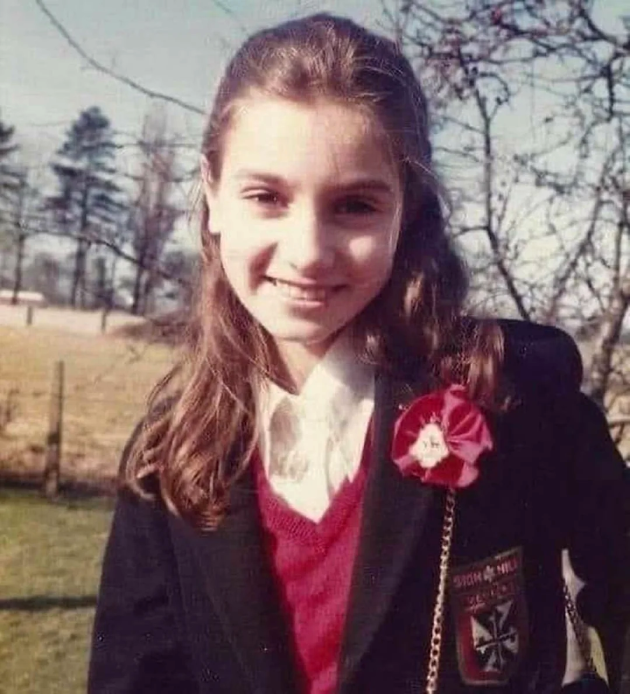 A young girl with long brown hair smiles outdoors, wearing a black blazer with a crest, a red sweater, and a white shirt. She has a red flower pinned to her blazer. Trees and a field are visible in the background.