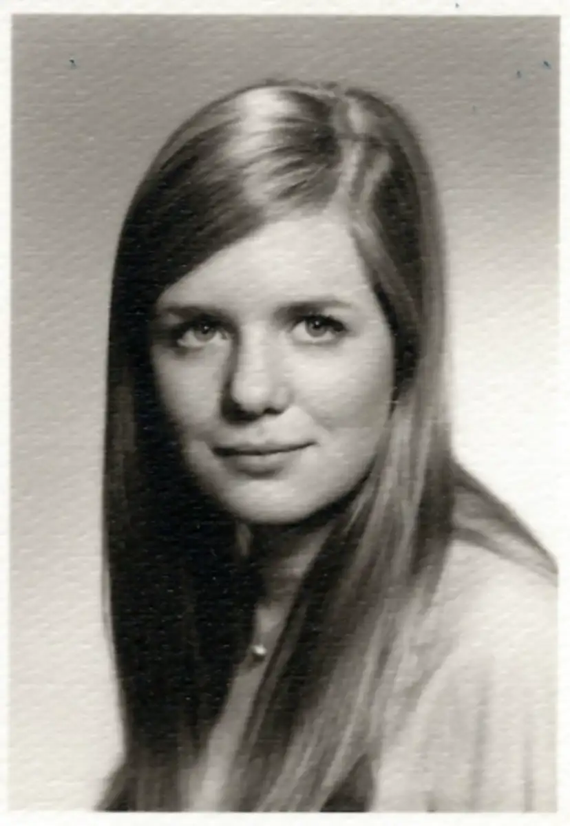 A black-and-white portrait of a young woman with long, straight hair, gazing slightly to the left of the camera. She has a neutral expression and is wearing a light-colored top. The background is plain.