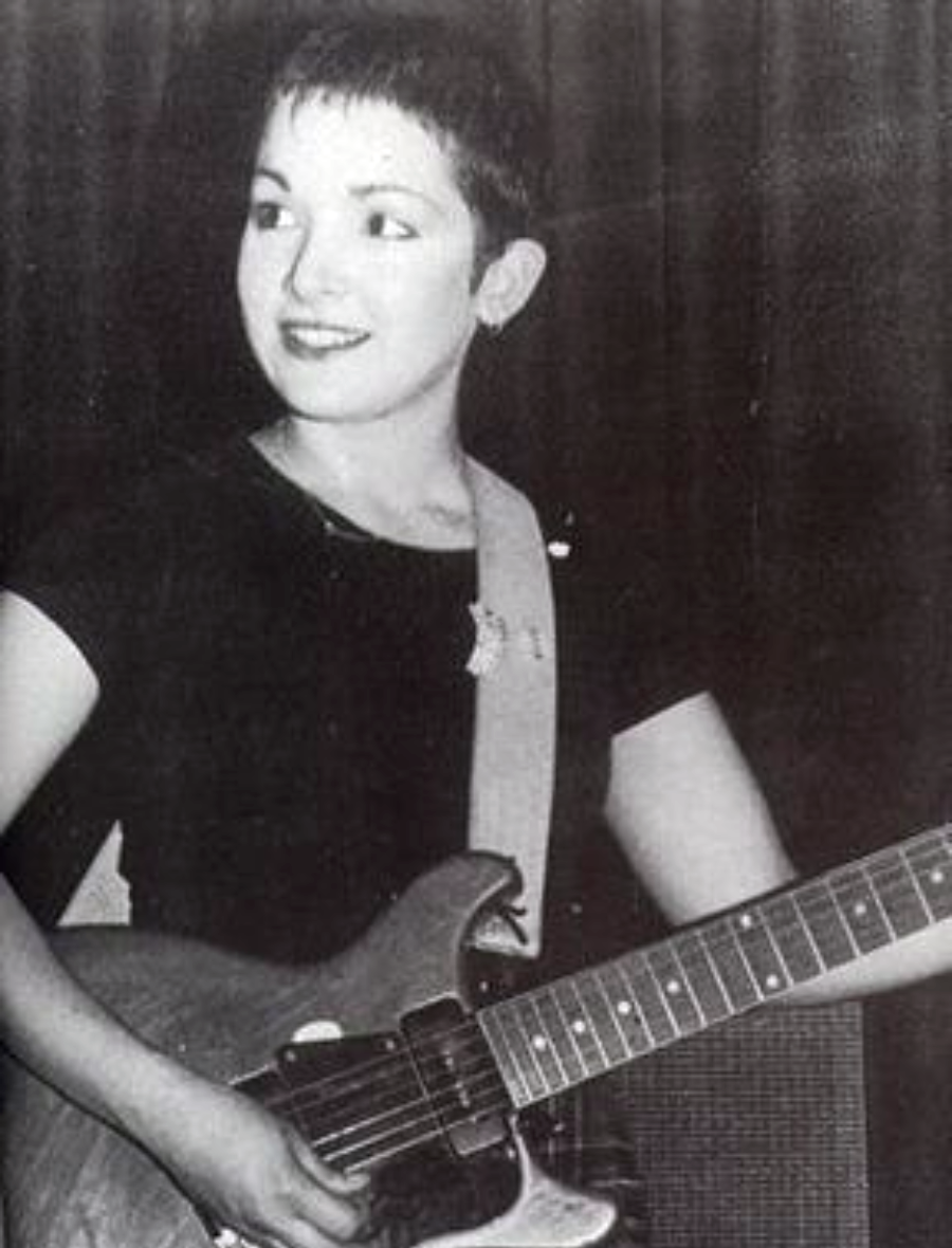 A young woman with short hair plays an electric guitar, smiling and looking to the side. She is wearing a dark shirt and standing in front of a dark background. The photo is black and white.