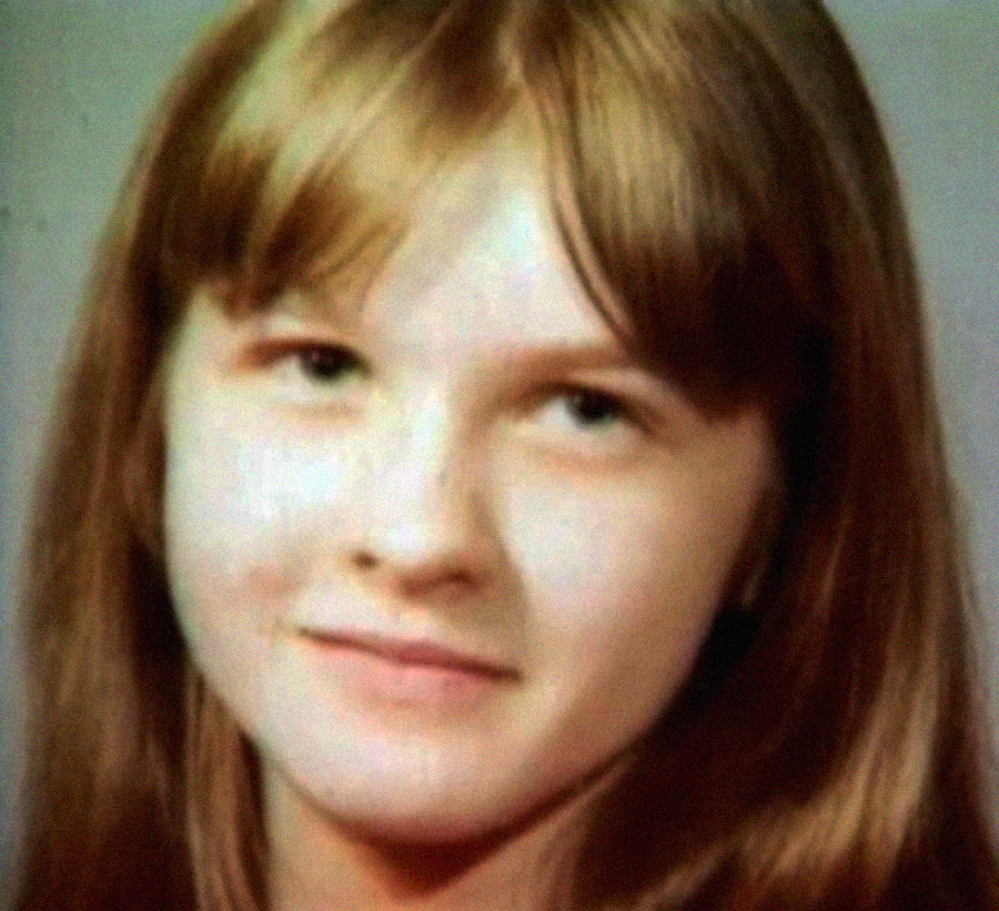 A close-up portrait of a young girl with long, straight light brown hair and bangs, looking slightly to the side with a neutral expression. The background is plain and softly blurred.