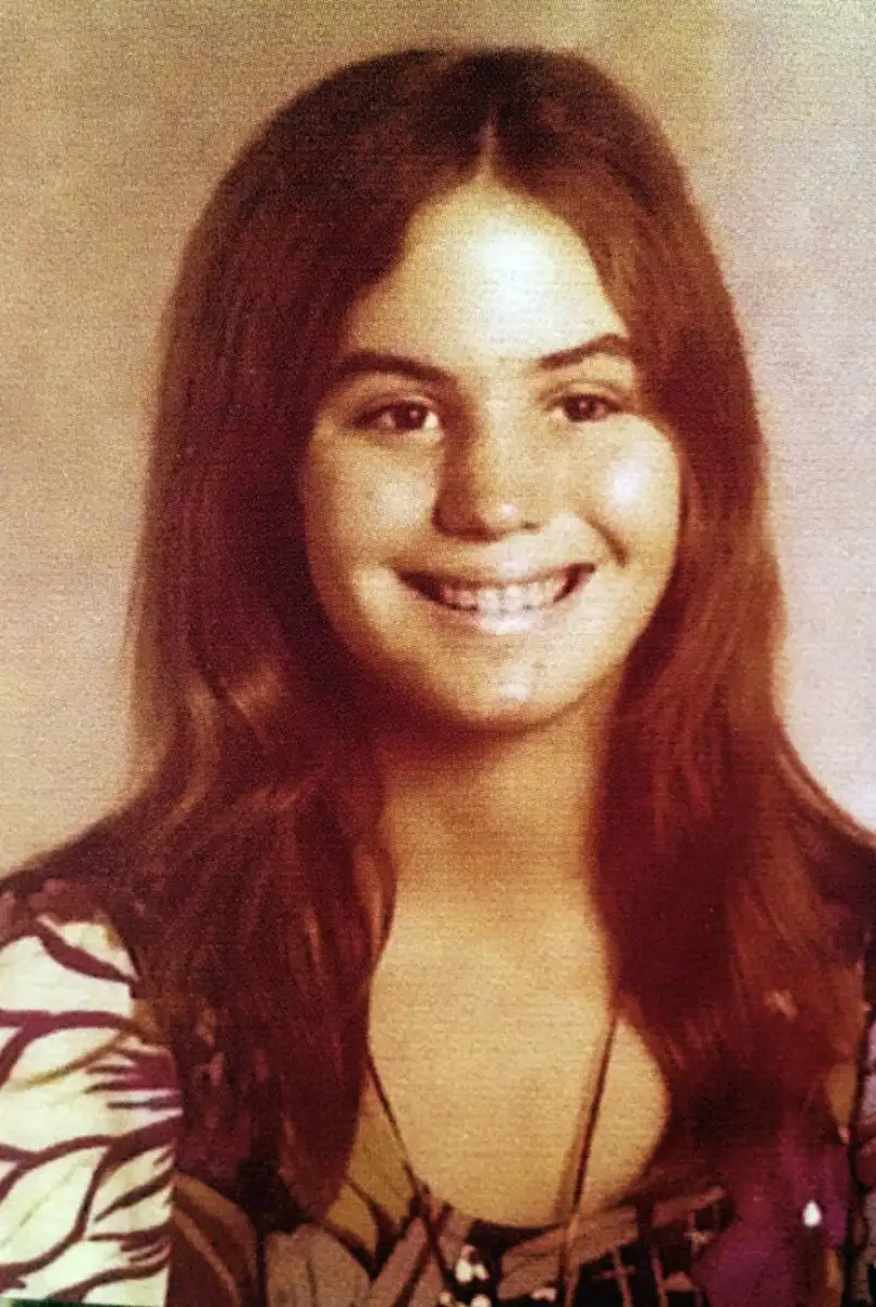 A young girl with long brown hair smiles at the camera. She is wearing a patterned top with purple and white designs and a necklace. The photo has a vintage, slightly faded appearance.