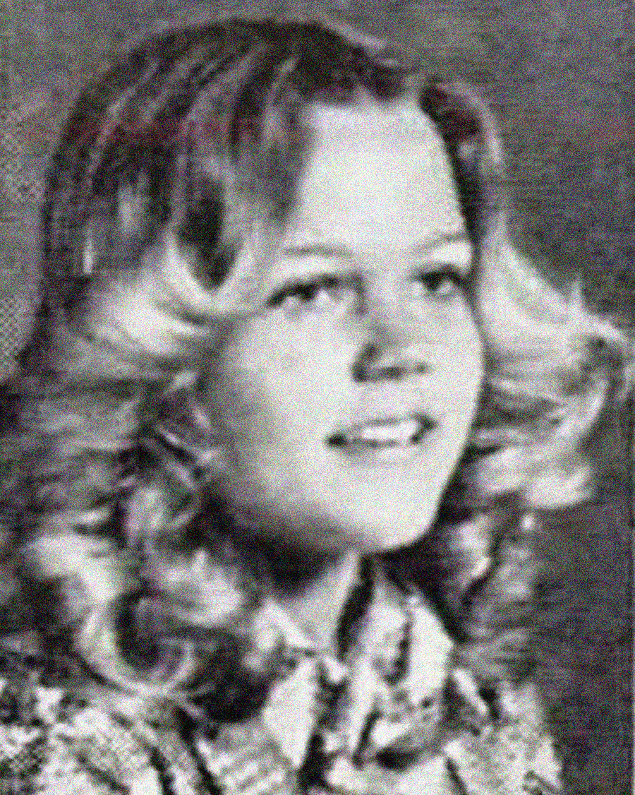 Black and white portrait of a young woman with shoulder-length wavy hair, wearing a patterned blouse, looking slightly to the side and smiling. The image has a vintage, yearbook-like quality.