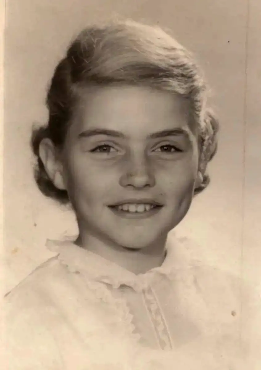 A black-and-white portrait of a young girl with light hair styled back, smiling and showing dimples, dressed in a light-colored, ruffled collared blouse. The photo has a vintage appearance.