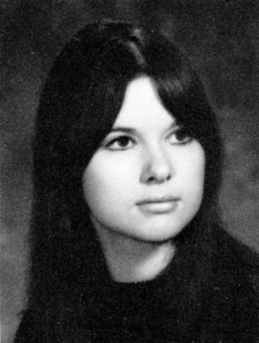 Black and white portrait of a young woman with long dark hair, looking slightly to her left. She has a calm expression and is wearing a dark top, with a plain background behind her.