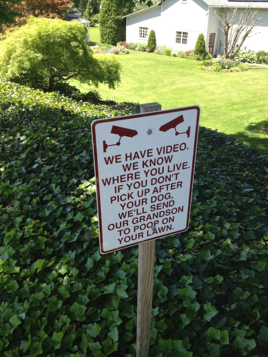 A sign in front of a house with security camera icons reads: "We have video. We know where you live. If you don't pick up after your dog, we'll send our grandson to poop on your lawn.
