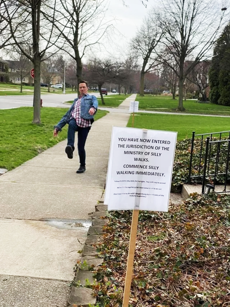 A man on a sidewalk lifts one leg high while walking past a sign that says, “You have now entered the jurisdiction of the Ministry of Silly Walks. Commence silly walking immediately.”