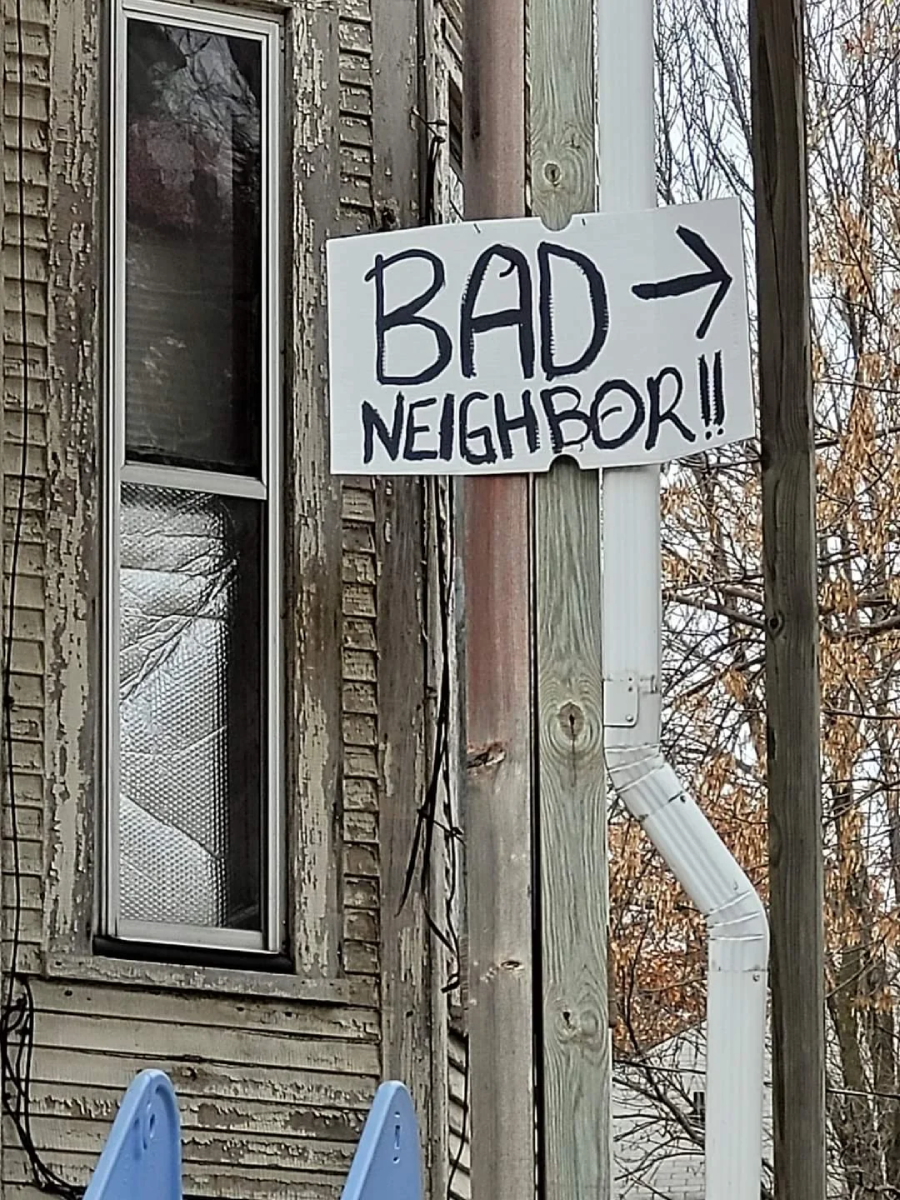 A handwritten sign reading "BAD NEIGHBOR!!" with an arrow points toward a neighboring house, attached to a weathered wooden post beside an old window. Trees and siding are visible in the background.