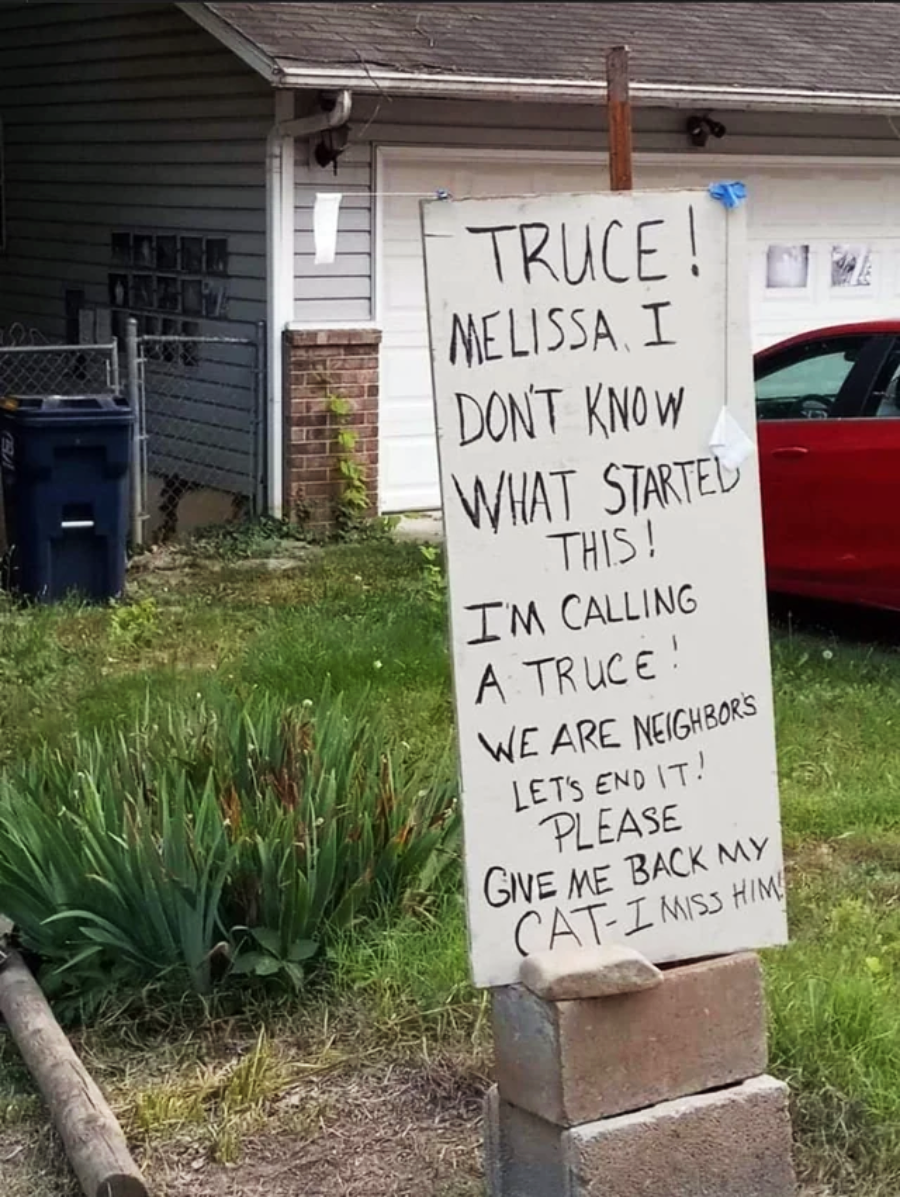 A handwritten sign in a yard reads: "TRUCE! Melissa, I don’t know what started this! I’m calling a truce! We are neighbors, let’s end it! Please give me back my cat. I miss him.