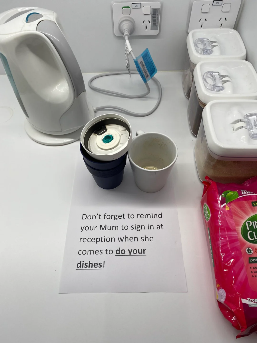 A countertop with a white electric kettle, two cups, three containers, and a packet of wipes. A sign reads: "Don’t forget to remind your Mum to sign in at reception when she comes to do your dishes!