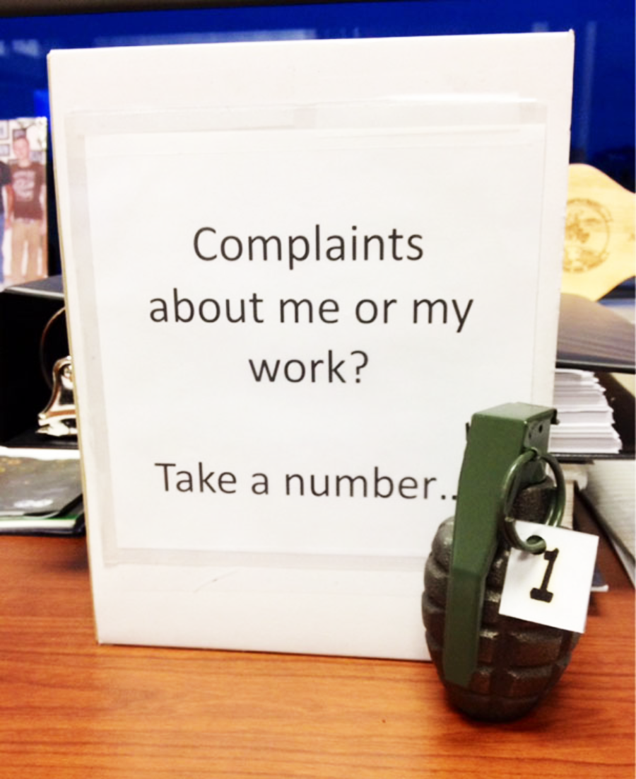 A sign reads "Complaints about me or my work? Take a number." In front of the sign is a replica hand grenade with a numbered tag "1" attached to its pin, suggesting humorously that complaints are risky.