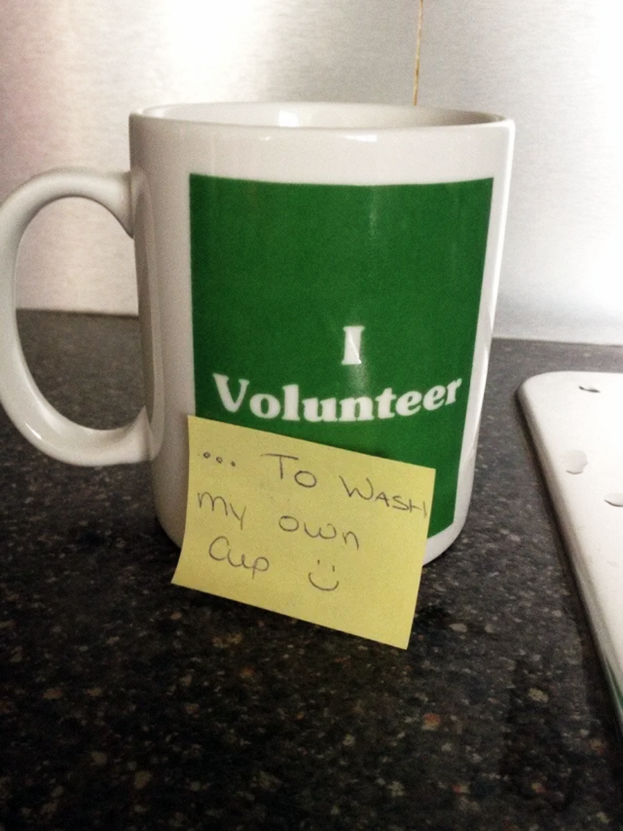 A white mug with a green label that reads "I Volunteer" sits on a dark countertop. A yellow sticky note on the mug says “…To wash my own cup :)” handwritten with a smiley face.