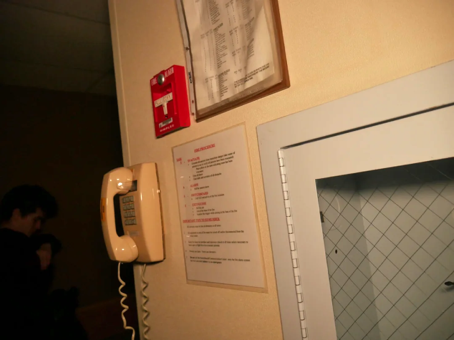 A beige wall-mounted corded phone is next to a fire alarm, a safety instruction sheet, and a metal panel with a wire-reinforced glass window. A person stands in a dim area in the background.