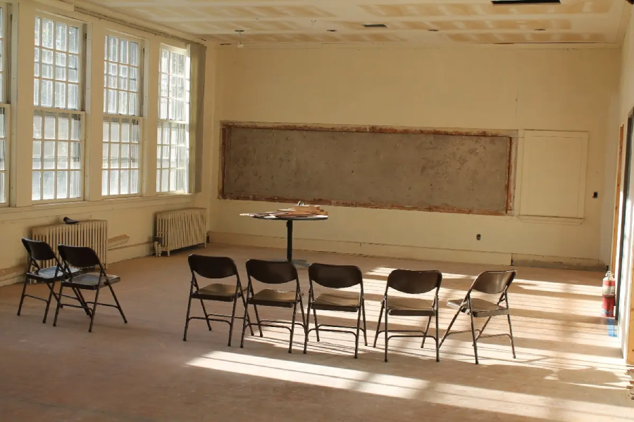 A nearly empty classroom with six folding chairs in a row, a small table with papers, large windows letting in sunlight, and a chalkboard with no writing on it.