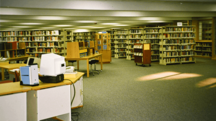 A spacious library with rows of bookshelves, desks with computers and office equipment in the foreground, and a book cart near the center, all under fluorescent lights.
