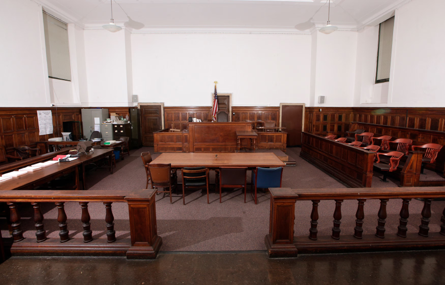 An empty courtroom with wooden benches, a judge’s bench, jury box, and desks for attorneys. An American flag stands behind the judge's bench, and there are chairs and office supplies throughout the room.