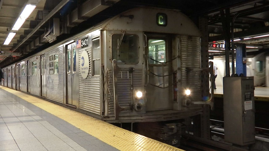 A silver subway train with the letter "C" on its sign stops at an underground station with yellow tactile paving and tiled floors. A few passengers stand on the platform.