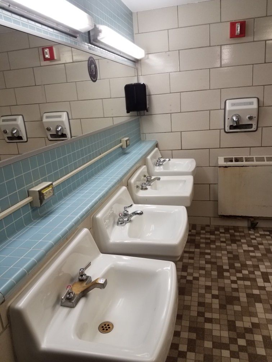 Four white sinks with silver faucets are lined up in a public restroom with tiled walls and floor; two hand dryers, a black soap dispenser, a mirror, and fluorescent lights are visible above the sinks.