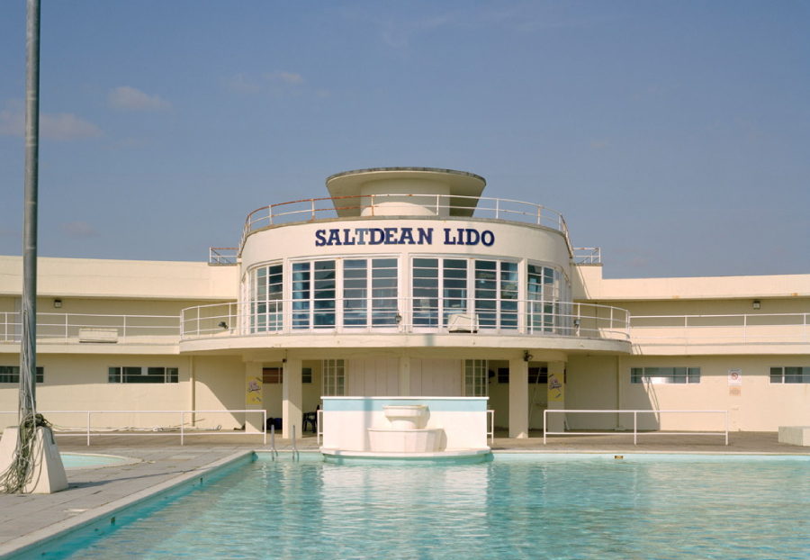 A view of Saltdean Lido, featuring a white, curved Art Deco building with large windows, a rooftop deck, and a pool with a central fountain in the foreground, under a clear blue sky.
